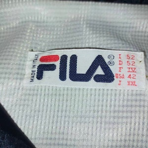 May include: Close-up of a white label with the FILA logo in navy blue and red. The label includes text that reads "MADE IN ITALY" and size information: I 52, D 52, F IV, USA 42, J XXL. The label is on a white and navy blue fabric.