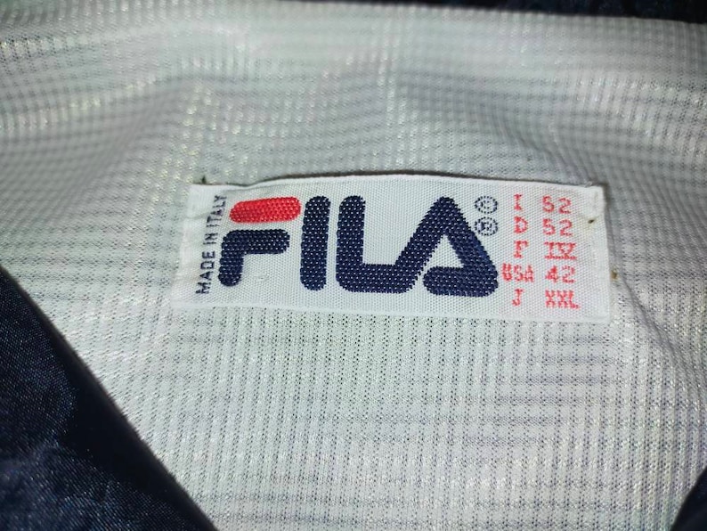 May include: Close-up of a white label with the FILA logo in navy blue and red. The label includes text that reads "MADE IN ITALY" and size information: I 52, D 52, F IV, USA 42, J XXL. The label is on a white and navy blue fabric.