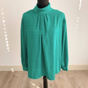 May include: A green, long-sleeved blouse with a high mock neck and a textured pattern. The blouse has a gathered detail at the neckline and a relaxed fit.