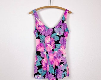May include: A black one-piece swimsuit with a floral print in shades of pink, purple, and teal. The swimsuit has a V-neck and a fitted bodice with a flared skirt.