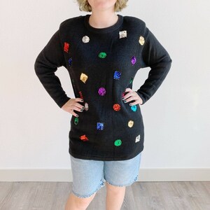 May include: Black long-sleeve sweater adorned with colorful sequins in various shapes. The sequins are in shades of red, gold, green, purple, and silver. The sweater has a crew neck and shoulder pads.