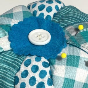 May include: A handmade pincushion with a blue felt flower and white button center. The pincushion is made of fabric in various patterns, including teal and white plaid, polka dots, and stripes. Yellow pins are inserted into the cushion.