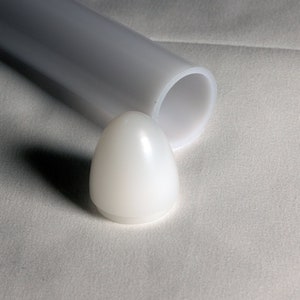 May include: A white, egg-shaped object with a flat base, next to a white cylindrical tube. The objects are made of a smooth, opaque material. The background is a light-colored, textured fabric.