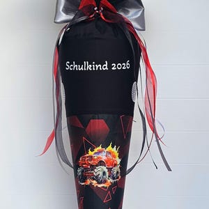 Può includere: Un cono scolastico nero e rosso con un motivo di monster truck e la scritta "Schulkind 2026". Il cono è decorato con un fiocco argentato e nastri rossi e neri. È sostenuto da un supporto metallico.