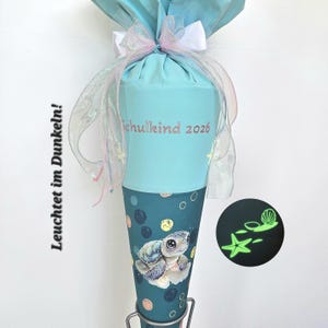 Può includere: Un cono scolastico turchese e blu con un'illustrazione di tartaruga e la scritta "Schulkind 2026". Il cono è decorato con un fiocco e un nastro. Il testo "Leuchtet im Dunkeln!" è sul lato.