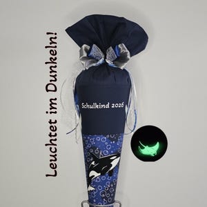 Op de afbeelding: Een marineblauwe schooltuit met een walvisontwerp en de tekst "Schulkind 2026". De tuit is versierd met een strik en linten. De tekst "Leuchtet im Dunkeln!" staat aan de linkerkant van de afbeelding.