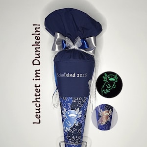 Könnte beinhalten: Eine marineblaue Schultüte aus Stoff mit weißem Axolotl-Motiv, Blasenmuster und dem Text 'Schulkind 2026'. Oben mit einer silber-blauen Schleife gebunden. Eine leuchtende Axolotl-Illustration ist in einem kreisförmigen Ausschnitt zu sehen.