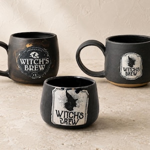 Puede incluir: Tres tazas de cerámica negro mate y un cuenco pequeño, cada uno con una etiqueta blanca con el texto "WITCH'S BREW" y la silueta de un sombrero de bruja. Los artículos tienen un acabado rústico y texturizado con tonos grises y oscuros.