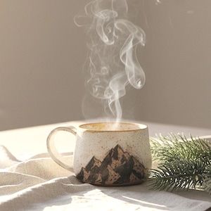 May include: A steaming mug with a mountain design sits on a table with a linen cloth. A sprig of evergreen and soft sunlight add to the cozy scene. The mug is beige with a brown mountain graphic.