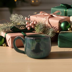 Könnte beinhalten: Eine Ansammlung von Geschenkboxen in Grün, Rosa und Creme, gebunden mit Bändern. Vor den Geschenken steht eine türkisfarbene Tasse, grüne Kugeln und Tannenzweige ergänzen das festliche Thema.