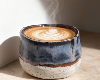 Könnte beinhalten: Eine Keramik-Kaffeetasse mit blau-weißer Glasur, gefüllt mit einem Latte. Die Latte-Art zeigt ein Blattmuster. Dampf steigt von dem heißen Getränk auf, was auf Frische hindeutet. Die Tasse steht auf einer hellen Oberfläche.