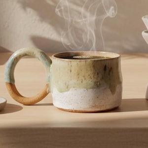 May include: A ceramic coffee mug with a large handle, filled with steaming coffee. The mug has a two-tone design, with a white base and a green and tan upper section. A small saucer with a coffee bean and a pour-over coffee maker are also present.