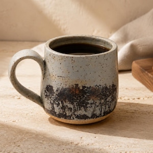 Può includere: Una tazza in ceramica grigio chiaro maculata con un design a silhouette di foresta marrone scuro attorno alla base. La tazza presenta un manico curvo e un bordo liscio e arrotondato, contiene un liquido scuro ed è posta accanto a un biscotto.