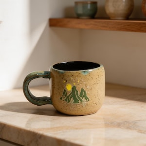 May include: A speckled tan ceramic mug with a dark interior and a curved handle. The exterior features a hand-painted green mountain landscape with a yellow moon and small yellow stars above the peaks, accented by small green cactus silhouettes.