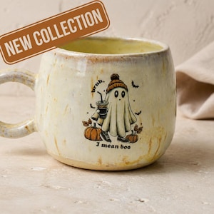May include: A cream-colored ceramic mug with a speckled finish featuring a graphic of a ghost wearing a beanie and sneakers, holding a coffee cup, surrounded by pumpkins and bats, with the text "bruh, I mean boo" printed below the ghost.