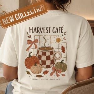 May include: An off-white t-shirt featuring a graphic on the back with the text "HARVEST CAFÉ" above a checkered mug, pumpkins, bows, and leaves. Below the graphic, the text reads "COFFEE • MATCHA • SLOW DAYS". A "NEW COLLECTION" banner is visible.