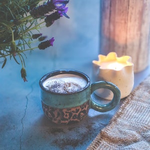 May include: A green and brown ceramic mug with a latte in it, a white candle with a flame, and a bunch of purple lavender flowers.