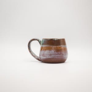 Puede incluir: Una taza de cerámica hecha a mano con un cuerpo redondeado y un asa grande. La taza presenta bandas horizontales de tonos marrón, naranja y morado, con un toque de verde azulado cerca del borde. La taza está diseñada para bebidas calientes.