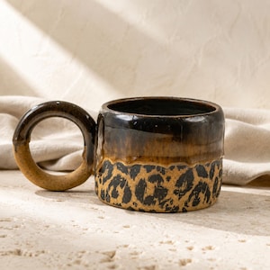 May include: A ceramic mug featuring a glossy dark brown upper section and a textured tan lower section with a black leopard print pattern. The mug has a large, circular, dark brown handle and a smooth, glazed finish throughout the exterior.