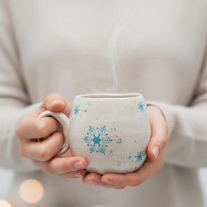 May include: A white ceramic mug with blue snowflake designs. Steam rises from the top of the mug, which is held by a person. The mug has a handle and is speckled with small dark spots.