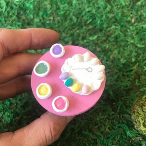 May include: A pink miniature table with a white plate and a small tool for decorating Easter eggs. There are four small bowls filled with different colored paints.