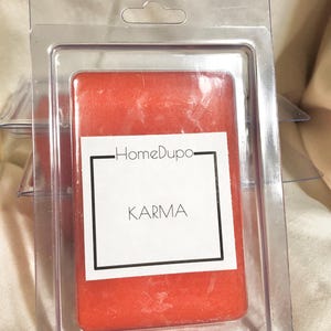 May include: A red wax melt bar in a clear plastic clamshell package. The label reads "KARMA" in a black frame, with "HomeDupo" above. The wax melt is a solid, rectangular shape, and the packaging is transparent.