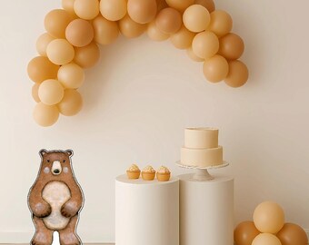 May include: A party scene featuring a brown bear cutout, a two-tiered cake, cupcakes, and a balloon arch in shades of brown and beige. The scene is set on white pedestals, creating a festive atmosphere.