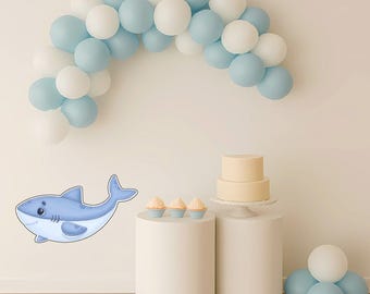 May include: A light blue cartoon shark graphic next to a white and light blue balloon arch. A two-tiered cake and cupcakes sit on white pedestals. Additional balloons are clustered on the floor.