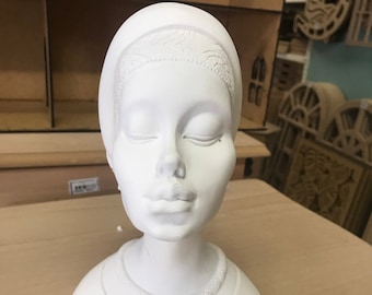 May include: A white bust of a woman with closed eyes. The sculpture has a head covering with a decorative band and a detailed neckline. The bust is on a light brown surface.