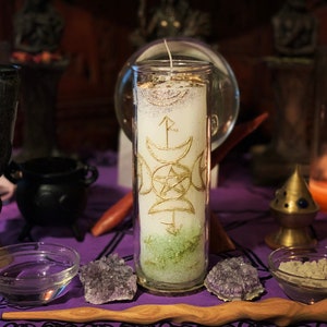 May include: A white candle with gold glitter and a gold design of a pentacle, triple moon, and other symbols. The candle is in a clear glass jar and is sitting on a purple table cloth.