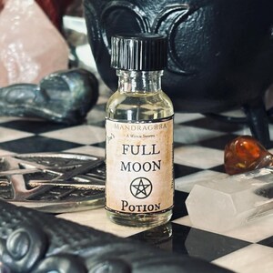 May include: A small glass bottle with a black cap and a label that reads "Mandrake - A Witch's Store" and "Full Moon Potion". The bottle is sitting on a black and white checkered surface with a silver pentagram and other crystals.