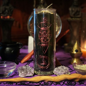 May include: A black candle with a pink and gold design in a glass jar. The candle is surrounded by crystals and other objects on a purple table cloth.