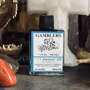 May include: A small, square glass bottle of "GAMBLERS" oil with a black cap. The label features a graphic of playing cards, dice, and a four-leaf clover. The bottle contains 1/2 fl. oz. (14.7ml) of oil and includes the text "7 Sisters Oil".