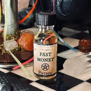 May include: A small glass bottle with a black cap and a label that reads "Mandragra A Witch Shoppe Fast Money Potion". The bottle is sitting on a black and white checkered surface.