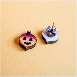 May include: A pair of wooden earrings shaped like a jack-o'-lantern. The earrings are painted in shades of orange, yellow, and purple. The jack-o'-lantern has a smiling face with black eyes and a wide mouth.