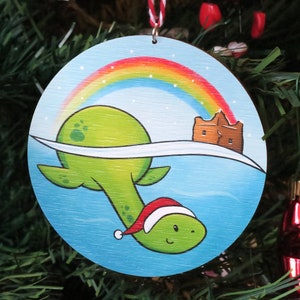 May include: A circular Christmas tree decoration depicting a cartoon Nessie wearing a Santa hat, swimming beneath a rainbow. The ornament has a light blue background with a small brown castle. A festive holiday decoration.
