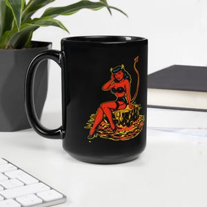 May include: Black ceramic mug featuring a retro-style illustration of a red devil woman with a tail and horns. The devil is sitting on a tree stump surrounded by flames. The mug has a large handle and is suitable for hot or cold beverages.