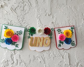 May include: A colorful paper banner with the word "UNO" in gold glitter letters. The banner is decorated with paper flowers in shades of yellow, pink, blue, white, and red.