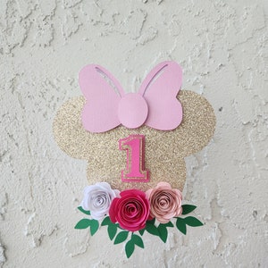 May include: Gold glitter Minnie Mouse cake topper with a pink bow, the number one, and pink, white, and peach paper flowers.