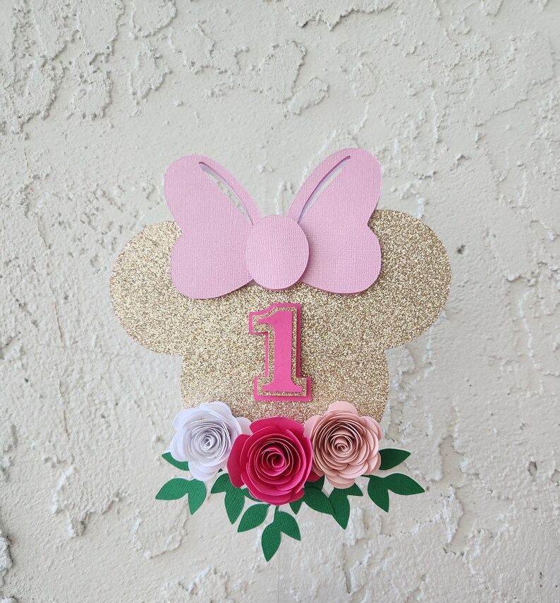 May include: Gold glitter Minnie Mouse cake topper with a pink bow, the number one, and pink, white, and peach paper flowers.