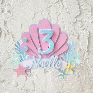 May include: A layered paper cake topper in a mermaid theme. The design features a pink seashell shape with a light blue number 3. The name "Noelle" is written in cursive on a banner. Starfish and sea plants complete the design.
