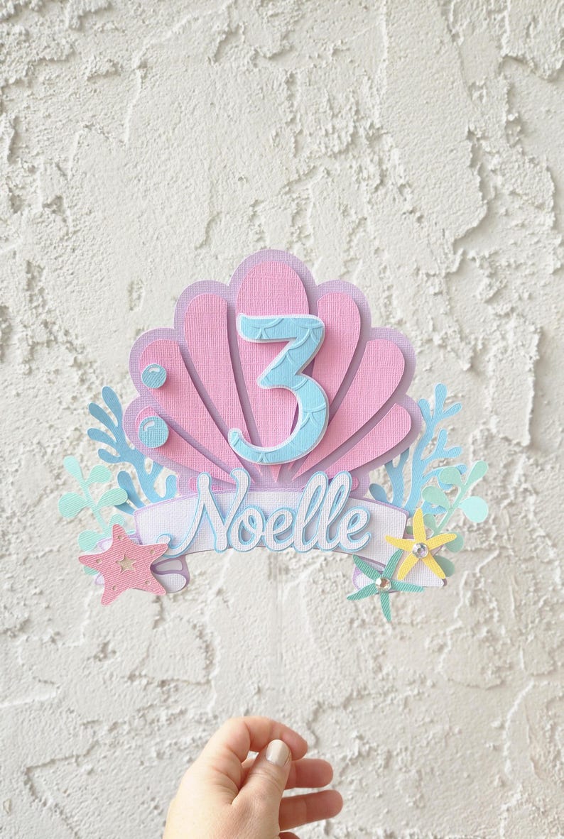 May include: A layered paper cake topper in a mermaid theme. The design features a pink seashell shape with a light blue number 3. The name "Noelle" is written in cursive on a banner. Starfish and sea plants complete the design.
