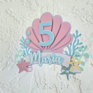 May include: A pink and blue mermaid-themed cake topper with the number 5 and the name "Maria". The design includes a seashell, starfish, and coral accents, perfect for a fifth birthday celebration.