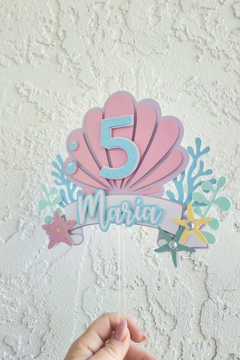 May include: A pink and blue mermaid-themed cake topper with the number 5 and the name "Maria". The design includes a seashell, starfish, and coral accents, perfect for a fifth birthday celebration.