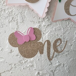 May include: Gold glitter cake topper with a pink bow and the word "one" in cursive. The topper is on a white stick.