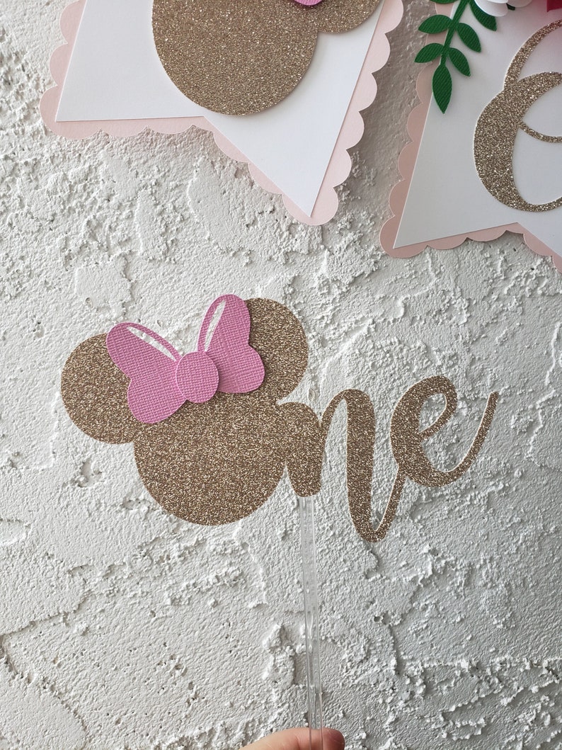 May include: Gold glitter cake topper with a pink bow and the word "one" in cursive. The topper is on a white stick.