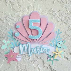May include: A layered paper cake topper with a seashell design in pink and lavender, featuring the number 5 in blue and the name "Maria". Includes starfish, bubbles, and aquatic plant accents. A festive decoration for a birthday celebration.
