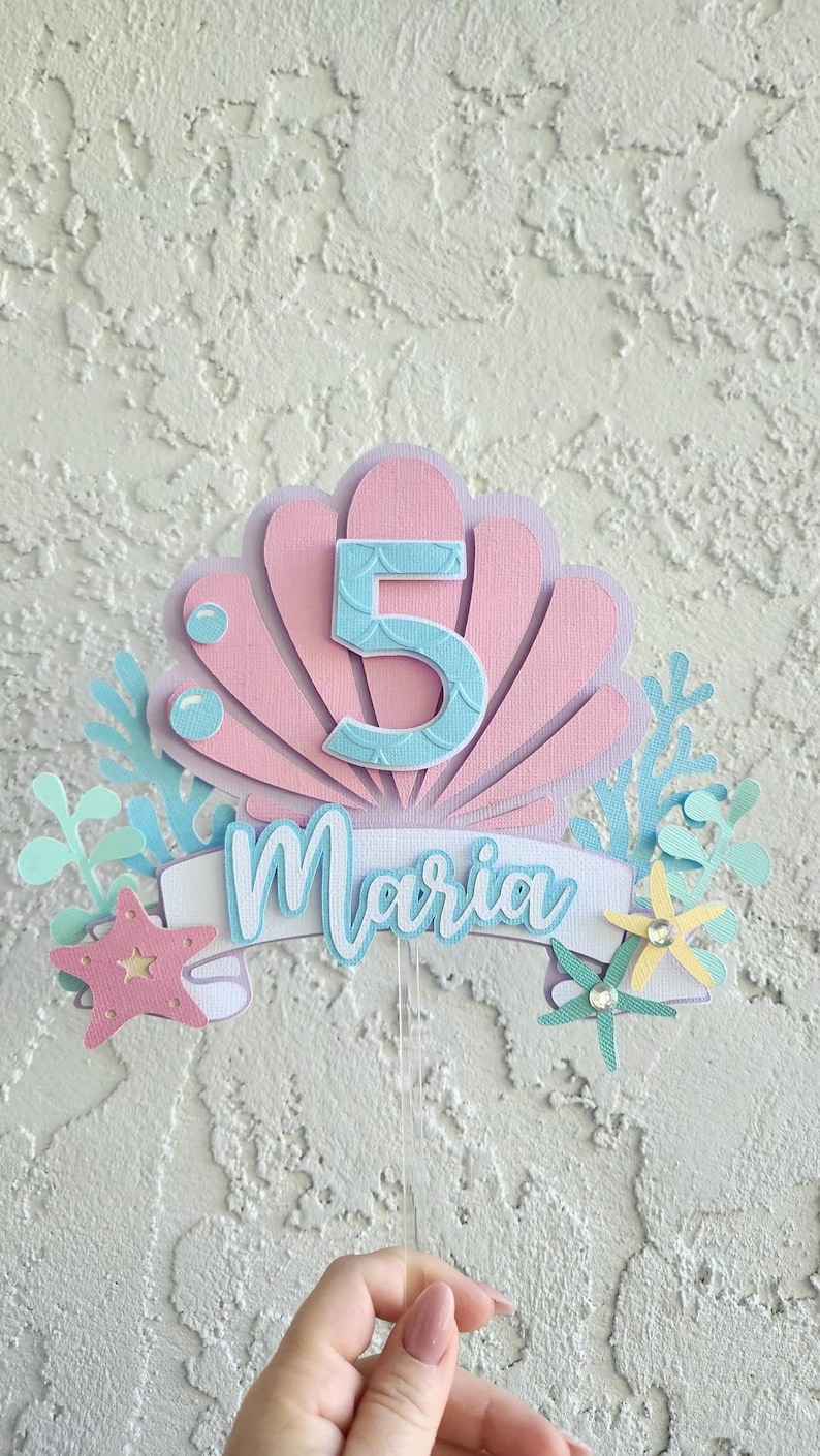 May include: A layered paper cake topper with a seashell design in pink and lavender, featuring the number 5 in blue and the name "Maria". Includes starfish, bubbles, and aquatic plant accents. A festive decoration for a birthday celebration.