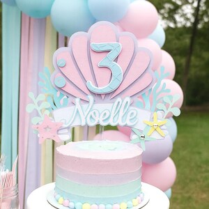 May include: A pastel-colored birthday cake with a seashell-themed topper that reads "Noelle" and the number "3". The cake is decorated with pastel frosting stripes and sits on a white cake stand. Balloons in shades of pink and blue are in the background.