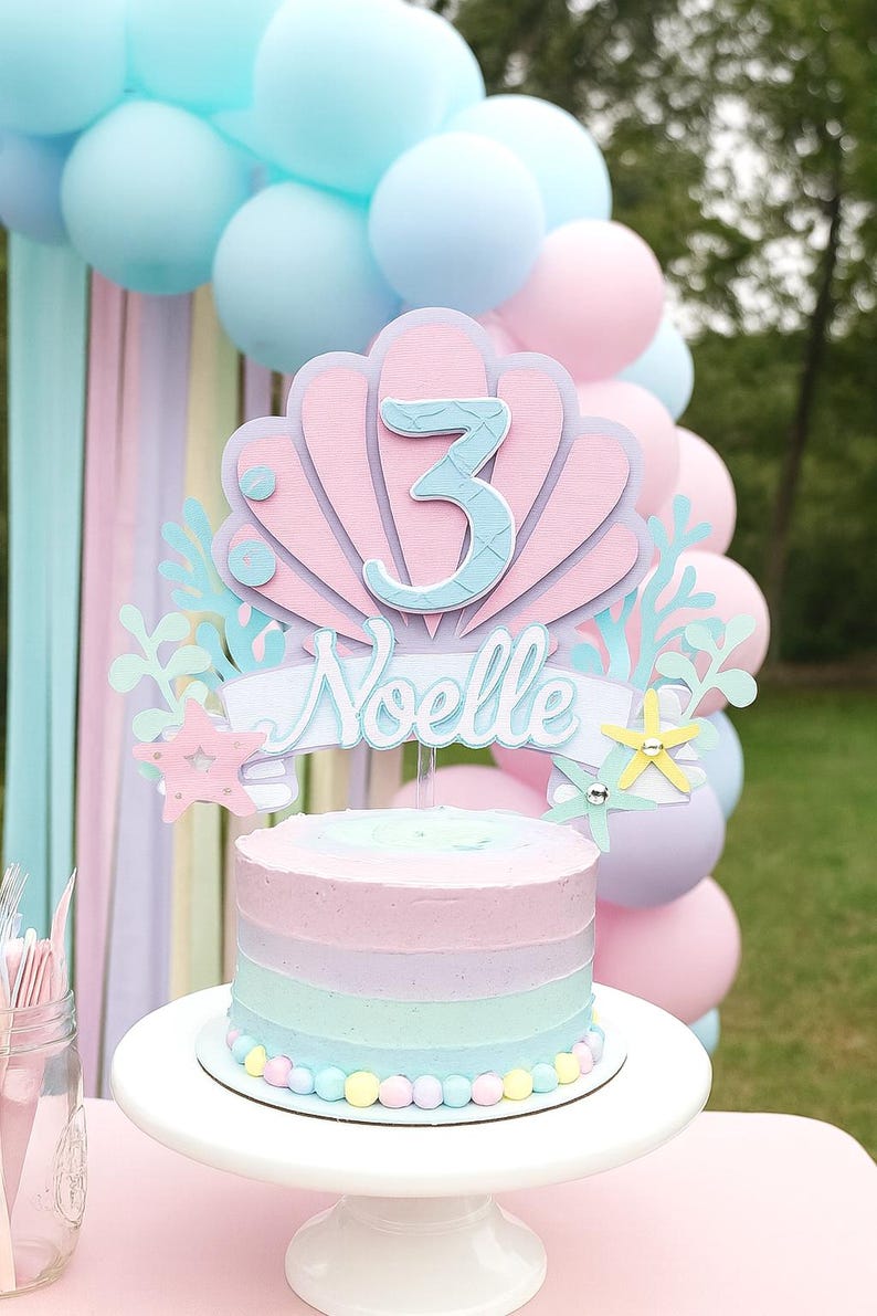 May include: A pastel-colored birthday cake with a seashell-themed topper that reads "Noelle" and the number "3". The cake is decorated with pastel frosting stripes and sits on a white cake stand. Balloons in shades of pink and blue are in the background.
